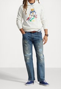 Vit hoodie med björngrafik och färgstark "Polo"-text, kombinerad med slitna ljusblå jeans och blå canvas-sneakers. Brun bälte och klocka.