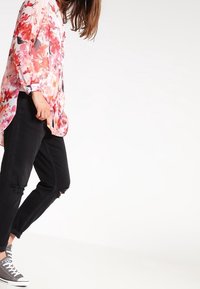 Blusa trasparente floreale con fiori rosa e rossi, maniche lunghe, abbinata a jeans neri strappati e sneakers grigie.
