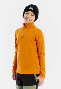 Pomarańczowy polarowy sweter z kołnierzem na pół zamek i logo na piersi; w zestawie z czarnymi i zielonymi spodniami narciarskimi oraz czarną dzianinową czapką.