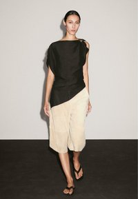 Haut en lin noir avec un design asymétrique ; culottes crème ; modèle portant des tongs noires ; arrière-plan minimaliste.