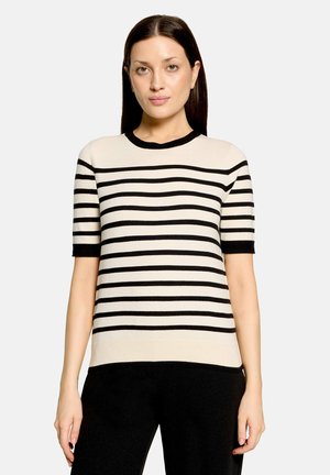 Femme aux cheveux lisses et foncés, portant un pull beige à manches courtes avec des rayures horizontales noires et un pantalon noir sur un fond uni.