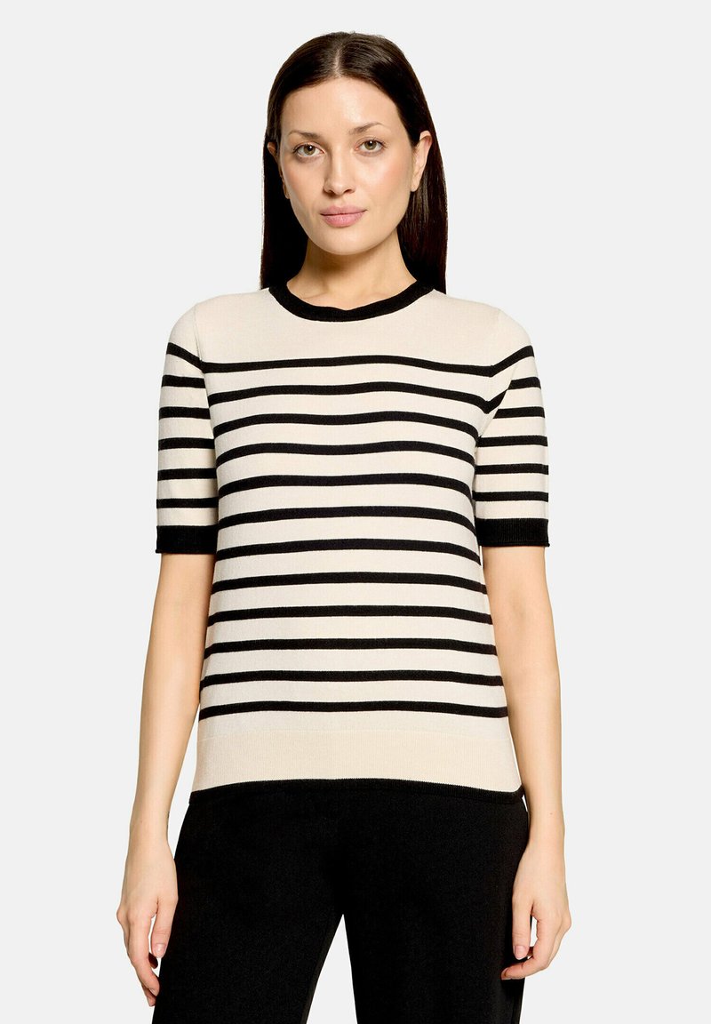 Femme aux cheveux lisses et foncés, portant un pull beige à manches courtes avec des rayures horizontales noires et un pantalon noir sur un fond uni.
