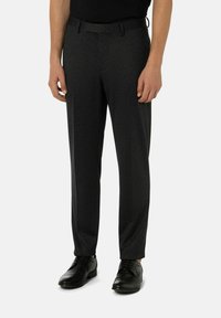 Homme portant un pantalon de costume à carreaux gris foncé, des chaussures en cuir noires et une chemise noire, se tenant contre un fond clair uni.
