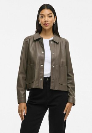 Veste courte en simili cuir marron avec un col, fermeture par bouton-pression et deux poches avant. Portée sur une chemise blanche et un pantalon noir.