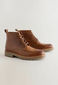 Brązowe skórzane buty za kostkę mają zaokrąglony czubek, sznurowany fason oraz teksturowaną gumową podeszwę z bieżnikiem zapewniającym przyczepność. Pętelka na pięcie.