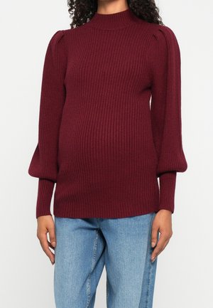 Femme portant un pull en maille côtelée bordeaux avec des manches longues bouffantes et un jean bleu sur un fond blanc uni.