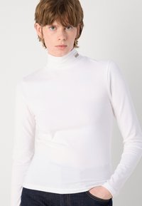 White long-sleeve turtleneck made of stretchy fabric, featuring a slim fit and a small branded tag on the neck. Paired with dark jeans.