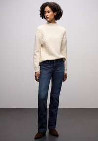Krémový svetr s pleteným vzorem, vysoký límec, volný střih, spárovaný s tmavomodrými rovnými džíny a hnědými semišovými kotníkovými botami. Jednoduchý, texturovaný design.