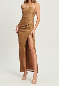 Złota sukienka bez ramiączek z dopasowanym gorsetem i bocznym rozcięciem. Posiada fakturowany materiał i gładką podszewkę. Przezroczyste obcasy dopełniają całość.