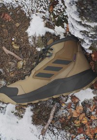 Brauner und schwarzer Wanderschuh mit "Cold.RDY" Text, der auf schneebedecktem Waldboden mit Blättern, Zweigen und einem moosbedeckten Stein in der Nähe ruht.