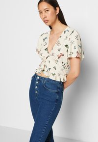 Blusa leve de creme com estampado de borboletas, bainha com babados e mangas curtas de sino, combinada com jeans de denim azul de cintura alta.