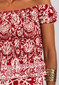 Top rojo sin hombros con patrones florales y de paisley en blanco, fruncido texturizado, dobladillo con volantes y mangas cortas abombadas. Pulseras metálicas doradas.