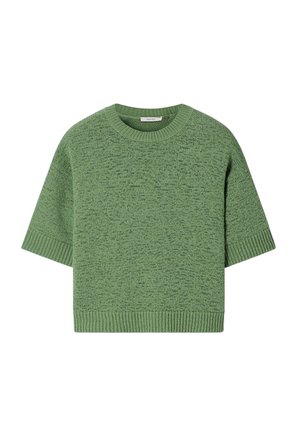 Vrouw met zonnebril, een losse groene gebreide top, lichtblauwe wijde jeans met kleine witte bloemenprints en beige geweven schoenen, poseert met opgeheven armen.