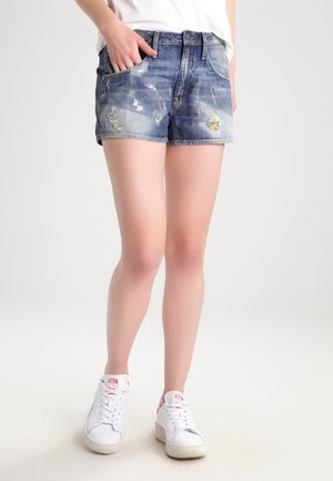 Vrouw die versleten blauwe denimshorts en witte sneakers draagt, staand met één hand in de zak tegen een effen achtergrond.
