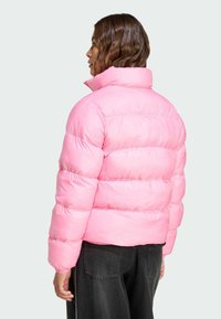 Een persoon met golvend haar draagt een felroze pufferjack, gezien van de achterkant tegen een lichte achtergrond. Donkere broek maakt de outfit af.