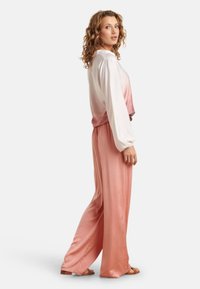 Lange, locker sitzende Hose aus zartem rosa Satin, kombiniert mit einem langärmeligen Oberteil in einem Weiß-zu-Rosa-Gradienten. Sanfte Textur und lässige Silhouette.