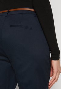 Navy trousers with a smooth texture, featuring a back pocket and a brown leather belt. The fabric appears tailored and fitted.