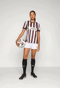 Futbola krekls ar brūnu un baltu svītrām, baltas bikses, melnas zeķes un melnas botas; tur rokās rakstainu futbola bumbu.