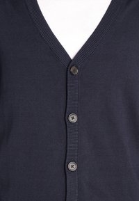 Navy sininen cardigan, joka on valmistettu teksturoituneesta kankaasta. Siinä on V-pääntie, ribattua viimeistelyä ja kuusi mustaa nappia edessä.