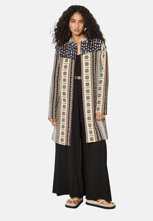 Frau mit langen lockigen Haaren trägt eine gemusterte lange Jacke über schwarzen, weit geschnittenen Hosen und beigen Sandalen und steht vor einem schlichten Hintergrund.