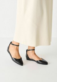 Ballerines en cuir noir avec un bout pointu, des côtés ouverts et une fine lanière à la cheville. Texture lisse avec un minimum de détails de couture.