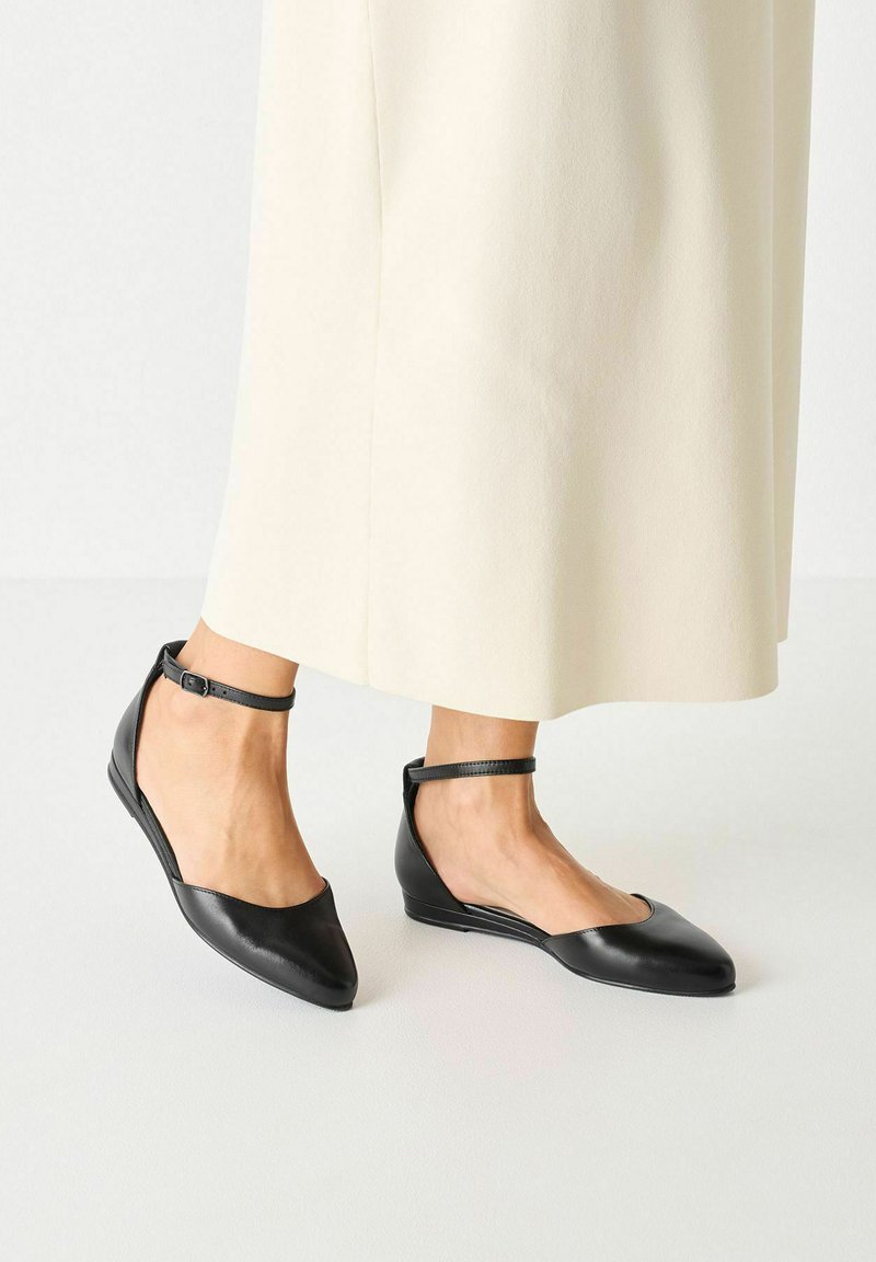 Ballerines en cuir noir avec un bout pointu, des côtés ouverts et une fine lanière à la cheville. Texture lisse avec un minimum de détails de couture.