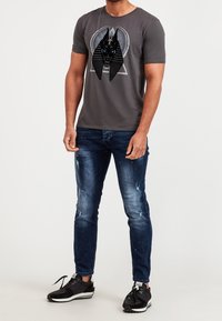 Grå kortärmad t-shirt med en svart grafik av ett hundhuvud med blå detaljer, i kombination med slitna blå jeans och svarta sneakers.