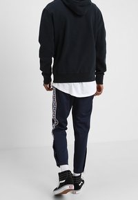 Felpa navy con dettagli a strisce laterali, felpa nera, camicia bianca a strati e sneakers nere. Il materiale sembra cotone, vestibilità comoda.