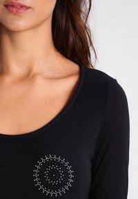 Svart långärmad skjorta med rund halsringning och ett dekorativt mönster av rhinestones på vänster sida av bröstet.