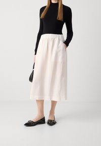 Donna con un top a maniche lunghe nero, gonna midi bianca con cintura elastica, ballerine nere a punta con fibbia, che porta una piccola borsa nera.