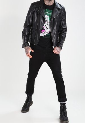 Schwarze Lederbikerjacke mit silbernen Reißverschlüssen, getragen über einem schwarzen T-Shirt mit grafischem, buntem Design. Kombiniert mit schwarzen Jeans und Kampfstiefeln.