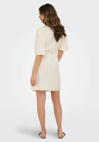 Abito beige corto con maniche a sbuffo, orlo all'altezza del ginocchio, tessuto leggero e un semplice design sul retro. Indossato con sandali piatti.