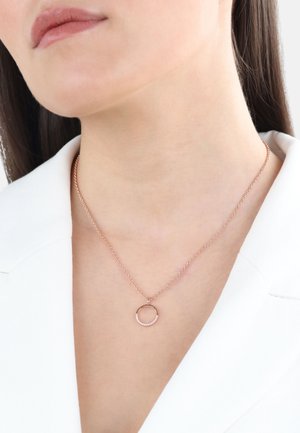 Collana in oro rosa con una delicata catena, caratterizzata da un pendente circolare decorato con piccoli cristalli. Indossata su una giacca bianca.