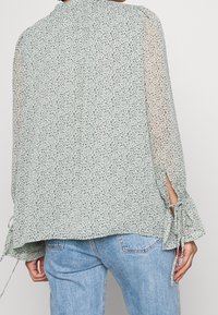 Ljusgrön blommig blus med långa ärmar, knytningar vid ärmsluten, mjukt genomskinligt tyg och en lös passform. Bakvyn visar en flödande silhuett.