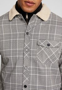 Grå och svart rutig skjorta med en krämfärgad fuskpälskrage, med knappar och en bröstficka. Strukturerad tyg med varierande mönster.