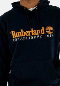 Sudadera azul marino con bolsillo tipo canguro en el frente, capucha con cordón y logo amarillo de Timberland con "ESTABLECIDO EN 1973" debajo.
