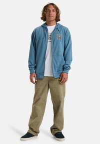 Light blue zip-up hoodie with a front pocket and logo detail, worn over a white t-shirt. Paired with loose tan pants and navy shoes.