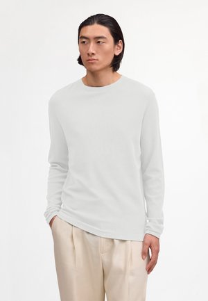 Mężczyzna model z czarnymi, średniej długości włosami, ubrany w prostą, białą koszulę z długim rękawem oraz beżowe spodnie, ręce częściowo schowane w kieszeniach.