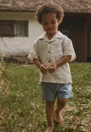 Mladé dieťa s kučeravými vlasmi, oblečené v béžovom tričku a rifľových kraťasoch, bosé, drží malé biele kvety, stojí na trávnatej zemi vonku.