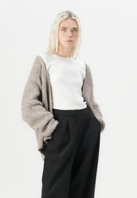 Světle hnědý pletený cardigan s volným střihem, nošený přes bílý top s kulatým výstřihem a černé kalhoty s vysokým pasem. Měkká textura s viditelnými stehy.