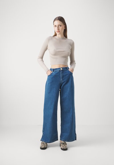 Haut beige à manches longues en maille côtelée associé à un pantalon en denim bleu foncé à jambes larges. La tenue comprend des poches avant et des chaussures à motifs.