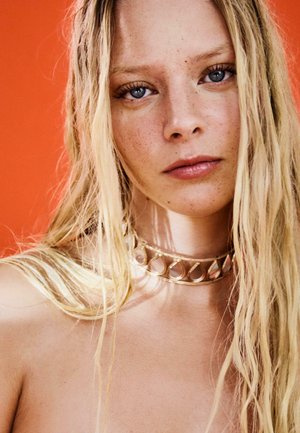 Jeune femme aux longs cheveux blonds, aux yeux bleus, avec des taches de rousseur et un collier doré en forme de goutte de larme sur un fond orange.