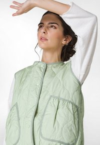 Hellgrüne gesteppte Weste mit runden Taschen, dekorativen Nähten und Druckknopfverschlüssen; über einem weißen Langarmshirt getragen.