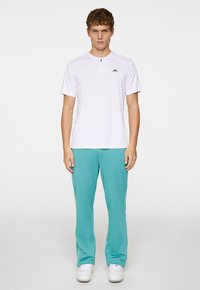 Camicia sportiva bianca a maniche corte con zip frontale e logo, abbinata a pantaloni sportivi turchesi con strisce bianche sui lati e dettagli con logo.
