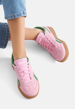 Różowe zamszowe trampki z zielonymi akcentami i białymi paskami, wyposażone w gumową podeszwę i wiązanie na sznurówki. Charakterystyczna, wyściełana językowa etykieta z logo.