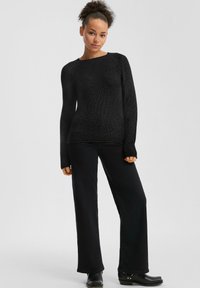 Zwarte gebreide trui met een geribbelde textuur, wijde halslijn en lange mouwen, gecombineerd met zwarte broek met wijd uitlopende pijpen en enkellaarsjes.