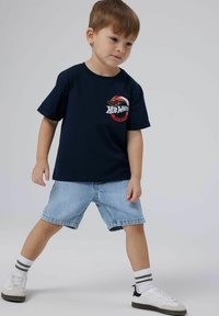 Tumši zils T-kreklis ar sarkanu Hot Wheels grafiku, kopā ar gaišiem džinsu šortiem, baltiem un pelēkiem apaviem un svītrainām baltām zeķēm.
