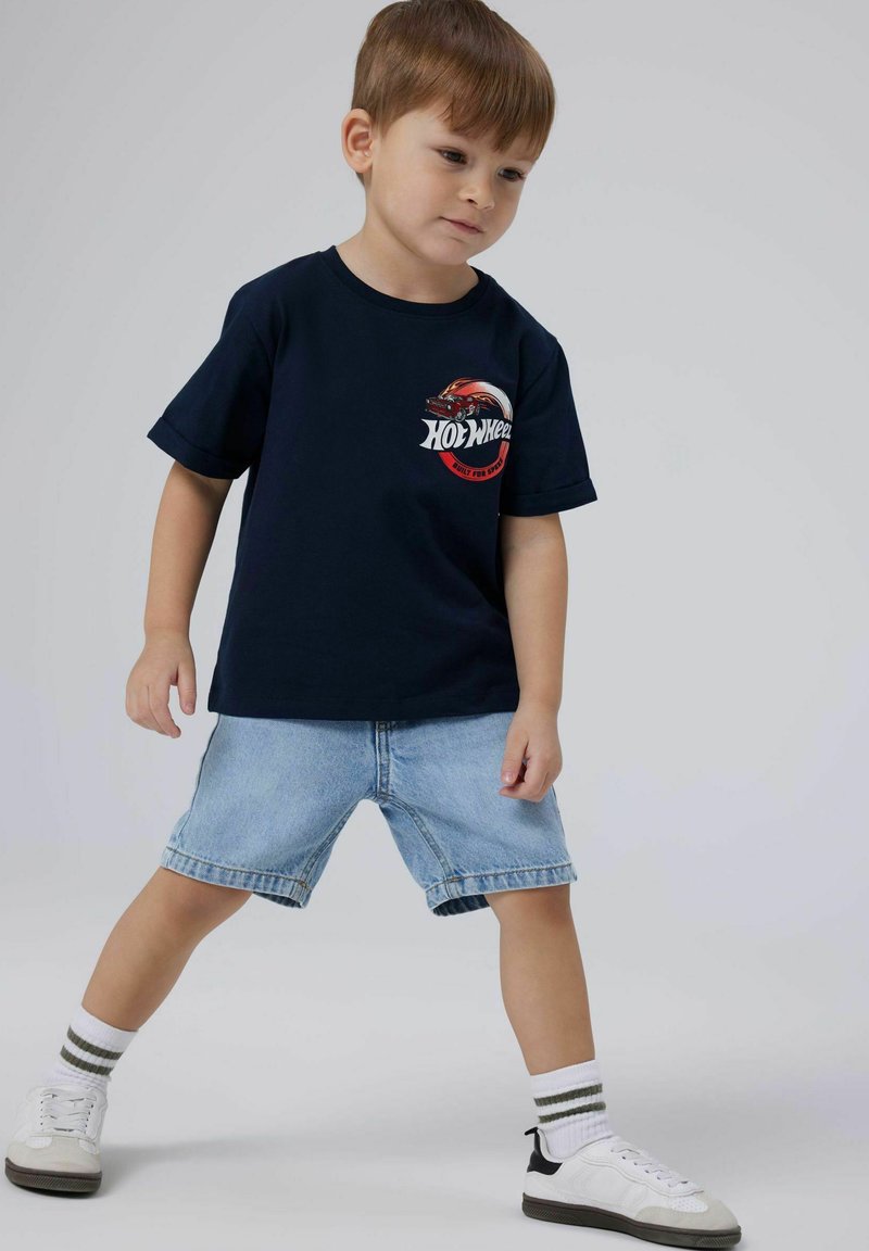 Tumši zils T-kreklis ar sarkanu Hot Wheels grafiku, kopā ar gaišiem džinsu šortiem, baltiem un pelēkiem apaviem un svītrainām baltām zeķēm.