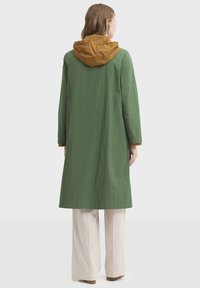 Imperméable vert avec une capuche marron, fabriqué en matériau léger. Caractérisé par des manches longues, une coupe droite et une longueur jusqu'aux genoux.