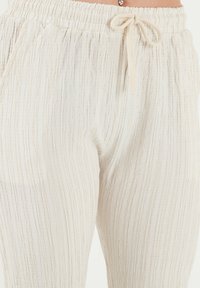 Lichtbeige elastische broek met verticale textuurstrepen, voorzien van een trekkoord en zijzakken. Zachte, lichtgewicht stof.